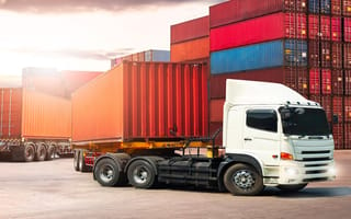 A tractor-trailer sits in an outdoor freight yard as a shipping container is loaded onto the truck bed.