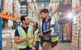 Two employees wearing bright green vests stand next to each other in a shipping warehouse, and one of them holds a tablet.