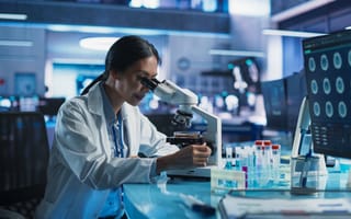 A woman wearing a white lab coat sits at a desk looking into a microscope.