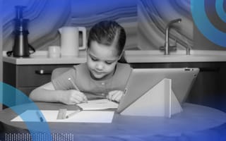 A child sitting at a table doing homework with an iPad propped up in front of her.