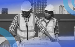 Two construction workers, one older and one younger, wearing safety vests and hard hats while looking over a blueprint together.