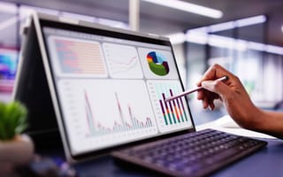 A close up of a hand holding a styles over a tablet screen that features a bar graph, pie chart and other data visualizations.