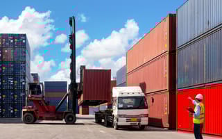 An employee wearing a hard hat and bright green vest stands in an outdoor freight yard directing a truck as it stacks shipping crates.