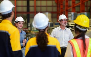 Five employees all wearing hardhats and other protective gear stand together in an industrial warehouse.