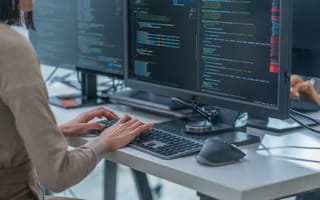 A close up of two hands typing on a desktop keyboard with lines of code displayed on the computer monitor.