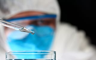 A scientist wearing a mask and other protective gear looks at a dropper as a small amount of liquid drips onto a circular petri dish.