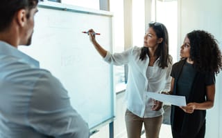 Two businesswomen analyzing a whiteboard and explaining the strategy to a man in the foreground.