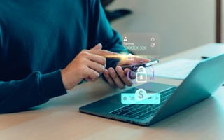 A close up of someone holding a mobile device over a laptop computer with a digital illustration featuring a dollar sign and padlock hovers in front to represent fintech.