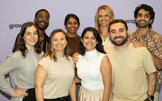 A group of Grow Therapy employees smile in front of a lavender wall printed with the company’s branding.