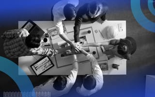 An aerial view of a team of six colleagues sitting at a table working on a project with their hands piled on top of one another’s in the center.