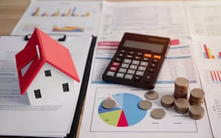 A calculator, stacks of coins and a small model of a house sit on top of financial documents filled with charts and graphs.