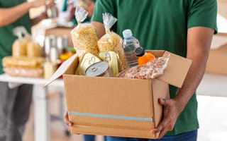Person carrying cardboard box filled with shelf-stable food items.