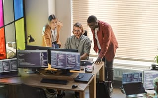 Three technologists analyze data together on a computer monitor screen in an office