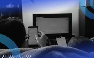 A view from behind of two people sitting on a couch, watching TV while also being on their smartphones.