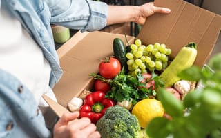 A close up of two hands opening a cardboard box filled with an assortment of green and red fruits and vegetables.