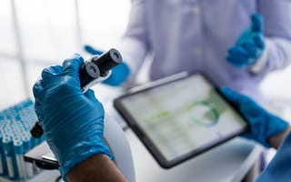 One hand wearing a blue glove adjusts a lab microscope while another hand wearing a blue glove holds a tablet device.