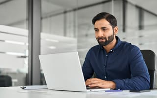 Busy Indian business man employee looking at laptop computer, businessman office worker or company manager analyzing project strategy working with ai software sitting at work desk.