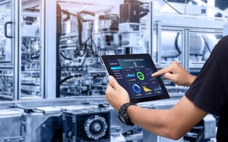 A pair of hands holds a tablet in front of a room full of industrial machinery.