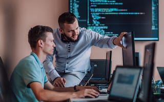 A software engineer points to a line of code on a computer monitor as he discusses an issue with a fellow software engineer seated next to him in an office.