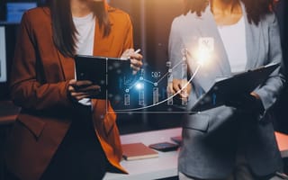 Two sales representative from the shoulders down compare notes and look at a light blue hologram of a positive graph.
