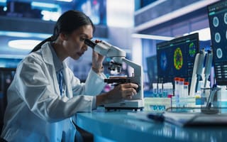 Biotech reseacher sits at a desk looking into a laboratory microscope.