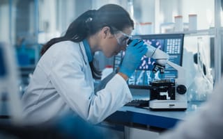 Scientist wearing a white coat sits at a desk looking into a lab microscope as she develops a biotech product.