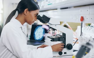 A biotech worker sits a desk in a lab looking into a microscope.