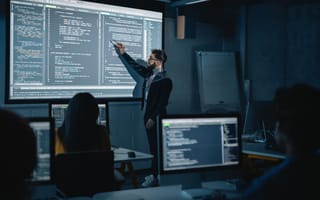 Professor stands in front of a class gesturing to a projector screen full of code to deliver a software lecture.