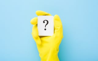 A yellow-gloved hand holds a white card with a black question mark against a solid light blue background.