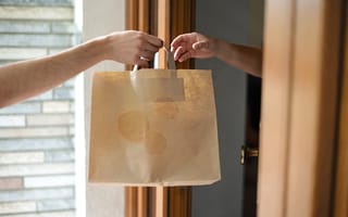 A close up of one person's hand passing a brown paper bag to another person's hand to represent food delivery.