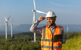 Worker wearing an orange vest and white hard hat stands in front of wind turbines.