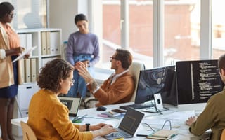 Two software engineers write code at a desk in an office while three of their coworkers discuss an ongoing project beside them