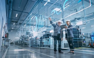 Two engineers in hard hats stand in a high-tech factory, engaging with virtual data displays projected in the air.