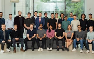 A diverse group of 25 people, casually dressed, pose smiling in an office setting with large windows revealing greenery outside, conveying a relaxed, team spirit.