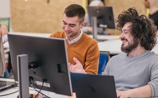 Two software engineers laugh while chatting together and working on their laptops in an office