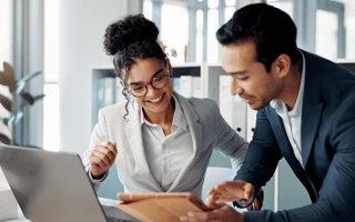 Two well-dressed professionals—a woman in a light gray blazer and glasses and a man in a dark suit—lean over a tablet together at a bright, modern office desk, with a laptop open nearby and shelves of binders in soft focus behind them.