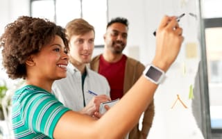 A software engineer writes notes on a whiteboard while two of her teammates watch on, with one taking notes in a notepad