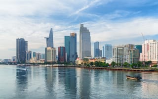 Modern city skyline of Ho Chi Minh City with diverse skyscrapers reflecting on calm river water, under a serene, partly cloudy sky.