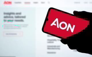 Hand holding a smartphone displaying the AON logo on a red background. The phone contrasts with a blurred computer screen in the background.