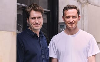 Two men smiling outside a building. One wears a dark shirt, the other a white t-shirt.