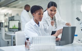 Two scientists in lab coats discuss data on a laptop in a bright lab.