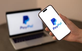 A hand holds a smartphone displaying the PayPal logo, with a blurred laptop showing the same logo in the background.