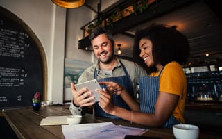 Two employees wearing aprons and holding a tablet device are pictured to depict a small business.