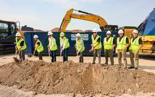 A line of people wearing green vests and hard hats at a construction site are pictured shoveling dirt.