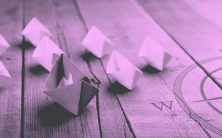 A paper boat leading a fleet of smaller boats on a wooden table with a compass image on it.