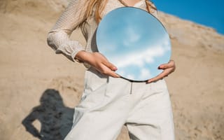 A woman in beige holds a mirror, reflecting the sky.