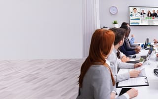 A group of people at a long conference table meet with a group of virtual team members.