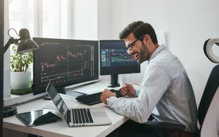 A happy white man sits at his desk with a laptop and two monitors that display trend charts.
