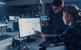 A designer and engineer consult a design on a computer monitor.