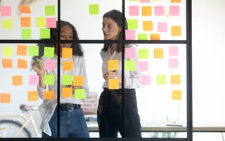 Image of coworkers discussing workflow laid out with sticky notes on a glass conference room wall.